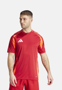 Maglia sportiva rossa con accenti arancioni, realizzata in materiale leggero. Presenta un collo a giro e maniche corte, logo sul petto.