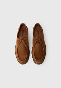 Scarpe in suede marrone con punta arrotondata, dettagli cuciti e lacci in pelle; suola piatta in gomma visibile. Nome del marchio all'interno.