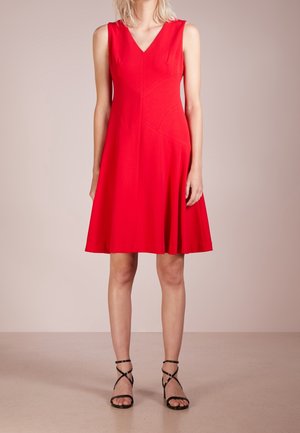Woman wearing sleeveless red knee-length dress with V-neck and black strappy high-heeled sandals, standing against neutral background.
