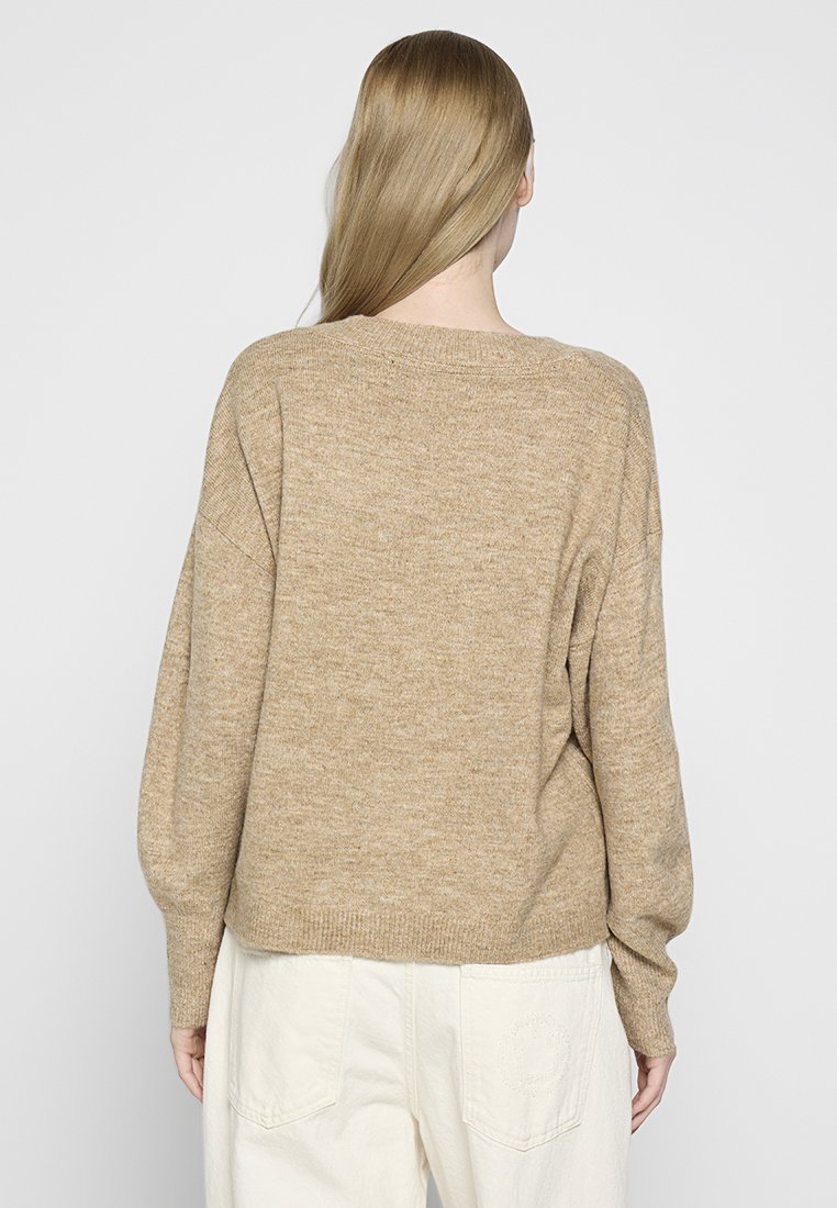 Mujer con cabello largo y rubio, vistiendo un suéter de punto suelto en tono beige y pantalones de color crema, de pie de espaldas contra un fondo claro y liso.