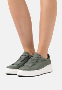 Green suede sneakers with a rounded toe, flat white rubber sole, and gray laces. Texture is soft and smooth, featuring subtle stitching details.