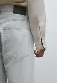 Lysgrå denimjeans har en baglomme og et sort lædermærke. En off-white langærmet skjorte med knapdetaljer er synlig.