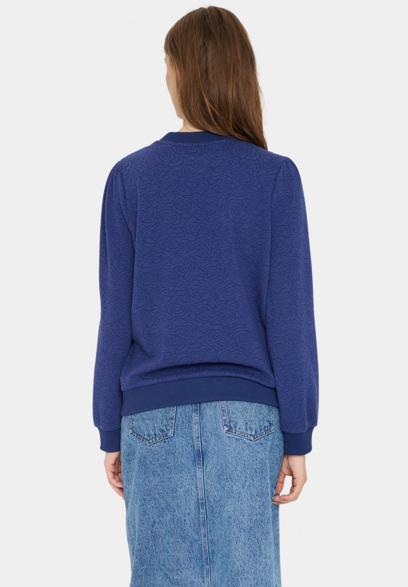 Blaues Sweatshirt mit einem strukturierten Rosenmuster, gerippten Bündchen und Saum, kombiniert mit einem Jeansrock. Die Rückansicht betont die lockere Passform und das Design.