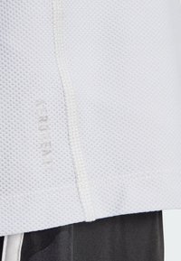 Camiseta atlética blanca hecha de tejido de malla texturizada, con una costura blanca y el logo "AEROREADY" cerca del dobladillo.