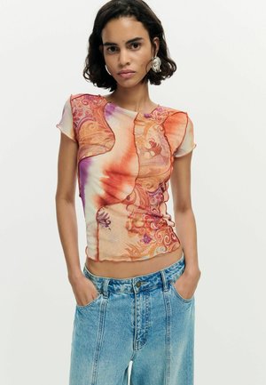 Žena s krátkymi tmavými vlasmi, oblečená v priliehavom oranžovo-fialovom vzorovanom tričku s krátkym rukávom a s vysokým pásom modrých džínsov, s rukami vreckách.