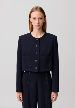 Donna con capelli biondi lisci che indossa una giacca corta nera abbottonata con due tasche frontali e pantaloni coordinati a vita alta.