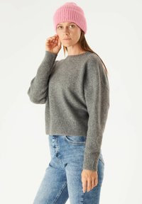 Grijze, cropped sweater met geribbelde manchetten en zoom, gecombineerd met blauwe, strakke jeans. Een lichtroze, geribbelde beanie wordt op het hoofd gedragen.