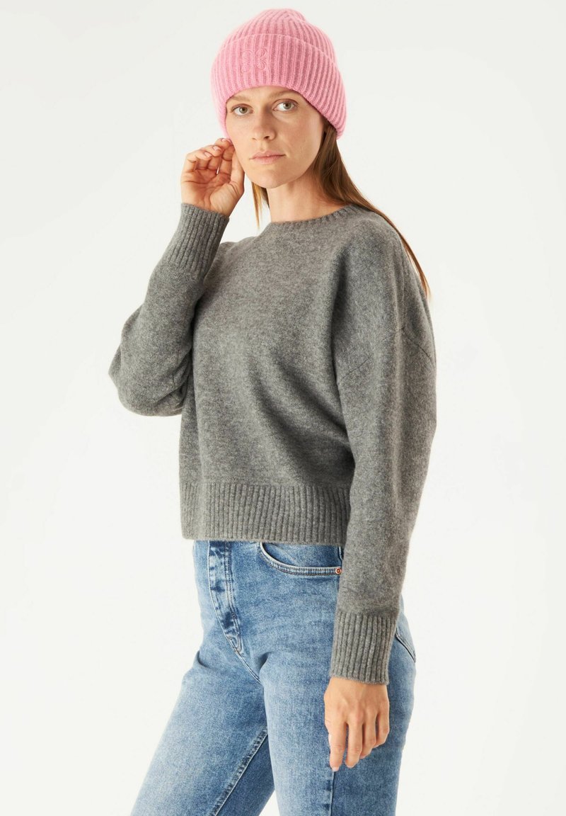 Grijze, cropped sweater met geribbelde manchetten en zoom, gecombineerd met blauwe, strakke jeans. Een lichtroze, geribbelde beanie wordt op het hoofd gedragen.