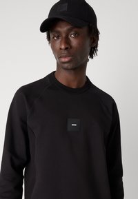 Μαύρο φούτερ με τετράγωνη ετικέτα λογοτύπου που έχει το κείμενο "BOSS". Ο σχεδιασμός περιλαμβάνει ραγκλάν μανίκια και rib κοπή στον λαιμό.