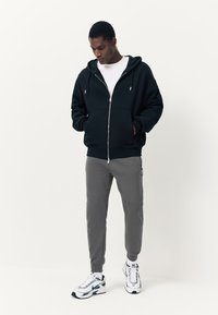 Black zip-up hoodie with front pockets and silver hardware, paired with gray tapered joggers. Model wearing white T-shirt and sneakers.
