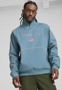 Gaiši zils džemperis ar augstu apkakli, rozā izšūts teksts "Definitely City" un atšķirīgu šuvju krāsa. Modelis valkā olīvzaļas bikses.