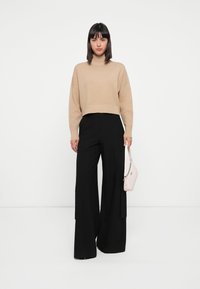 Pull beige court avec col roulé, associé à un pantalon noir large taille haute. Le mannequin tient un petit sac à main rose.