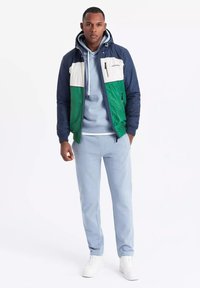 Färgblockerad huva-jacka i marinblått, grönt och kräm, kombinerad med en ljusblå hoodie och matchande joggerbyxor; vita sneakers.