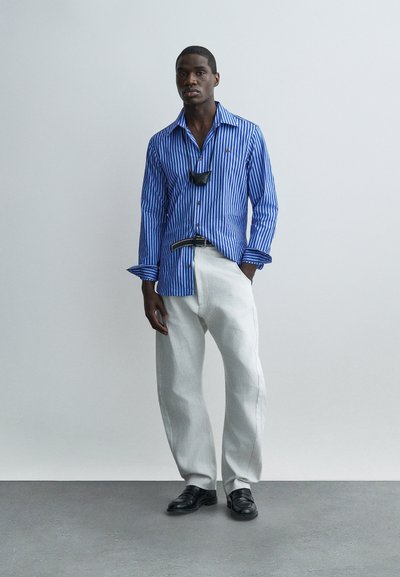 Άνδρας με μπλε ριγέ πουκάμισο, λευκό παντελόνι, μαύρη ζώνη, μαύρα παπούτσια και μαύρη μάσκα προσώπου κρεμασμένη στο λαιμό, στέκεται μπροστά σε απλό φόντο.