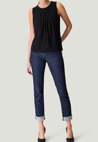 Frau in schwarzer ärmelloser Bluse, dunklen hochgekrempelten Jeans und schwarzen High Heels, stehend mit einer Hand hinter dem Rücken vor neutralem Hintergrund.