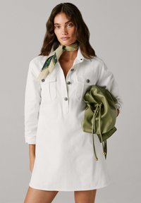 Abito camicia in denim bianco con chiusura frontale con bottoni, due tasche sul petto, maniche arrotolate e una borsa verde leggera con coulisse tenuta al braccio.