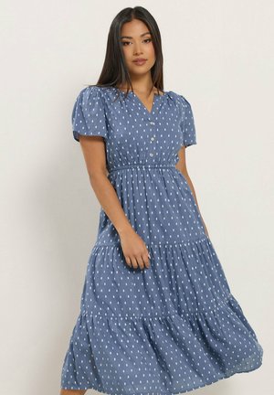 Vrouw draagt een blauwe, korte mouw, gelaagde jurk met een wit gestippeld patroon en een knoopsluiting in een V-hals, staand tegen een effen achtergrond.