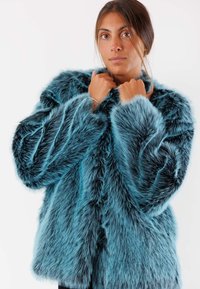 Giacca in pelliccia sintetica di colore teal, con una texture morbida, vestibilità ampia e maniche larghe. Il materiale presenta lunghezze variabili per un effetto di maggiore profondità.