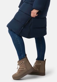 Brune ankelstøvler i tekstureret ruskind, med kontrastsnørebånd og en tyk gummisål. Båret med tætsiddende blå jeans og en navy pufferjakke.