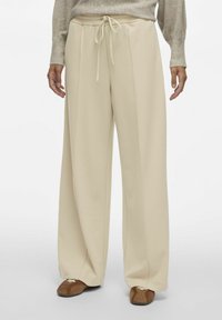 Breedbenige beige broek met een trekkoord in de taille, gemaakt van een zachte stof, met verticale naden en een relaxte pasvorm. Geaccentueerd met bruine schoenen.