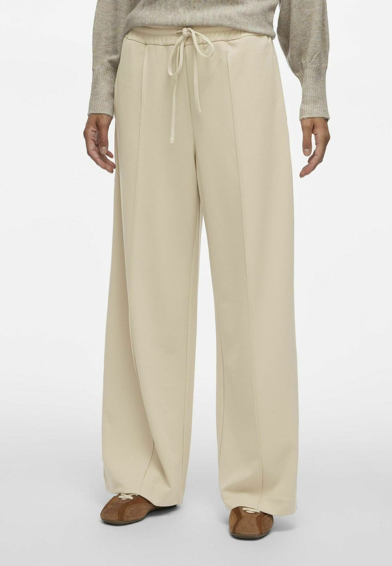 Breedbenige beige broek met een trekkoord in de taille, gemaakt van een zachte stof, met verticale naden en een relaxte pasvorm. Geaccentueerd met bruine schoenen.