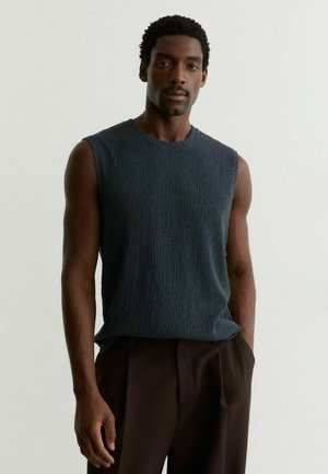 Mees, kellel on lühikesed lokkis juuksed, kannab tekstuurse pinnaga tumesinist varrukateta särki ja tumedaid pükse, seisab ühevärvilise heleda tausta ees.