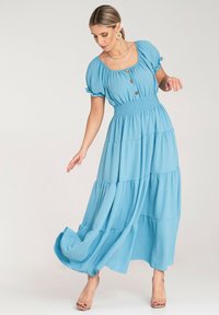 Niebieska maxi sukienka na poziomach z bufkami, z zapięciem na guziki przy dekolcie, elastycznym pasem, zwiewnym materiałem i lekką teksturą.