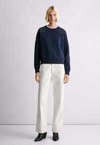 Felpa navy con dettagli di borchie sulle spalle, indossata con pantaloni bianchi a gamba larga e stivaletti neri con tacco. Texture liscia, vestibilità rilassata.