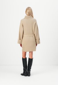 Manteau en tricot beige avec col montant, taille ceinturée et manches larges. Bottes noires montant jusqu'aux genoux visibles en dessous, présentant une finition texturée.