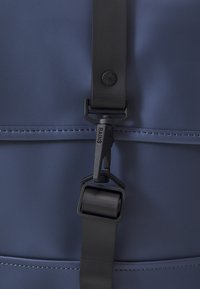 Zaino impermeabile navy con hardware nero opaco, tracolla regolabile e cuciture visibili lungo la cucitura superiore. Presenta un logo discreto.