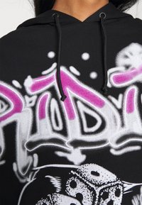 Sweat à capuche noir avec cordons noirs, arborant un texte style graffiti en blanc et rose ainsi qu'un motif de dés blancs en dessous.