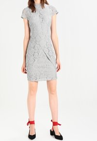 Harmaa pitsimekko lyhyillä hihoilla, tiukalla siluetilla ja sivurei'ällä, yhdistettynä mustiin korkokenkiin, joissa on punaiset nauha-akcentit.