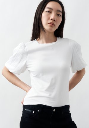 Sieviete ar gariem tumšiem matiem, valkājot baltu blūzi ar pūderpapēžu piedurknēm un melnas džinsas, pozu ar rokām uz gurniem pret vienkāršu fona sienu.