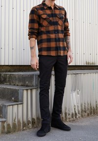 Ruudukko ruskea ja musta flanellikangas paita, jossa on kaksi rintataskua, yhdistetty mustiin slim-fit-housuihin ja mustiin lenkkareihin, seisoo betoniaskeleilla.