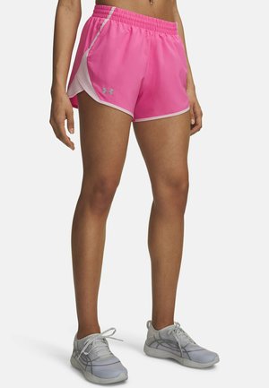 Mujer con pantalones cortos deportivos de color rosa brillante con ribete claro y zapatillas de correr grises, de pie contra un fondo claro y liso.
