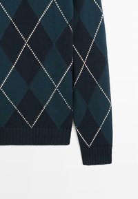 Donkergroene en marineblauwe argyle gebreide trui mouw met wit gestikt diamantpatroon en geribbelde boord op witte achtergrond.