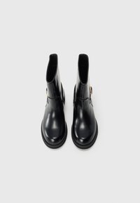 Black shiny leather ankle boots with round toes, flat soles, and gold buckle details on the side, viewed from above on a white background.