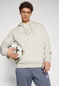 Beiger Hoodie mit Kängurutasche und Logodetail, kombiniert mit grauen Sweatpants und einem schwarz-weißen-grünen Fußball.