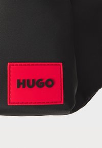 Juodas nailono krepšys su išsiskiriančiu raudonu guminiu logotipo pleistru, pažymėtu „HUGO“ juodomis raidėmis. Lygus paviršius su tvarkingais kampais.