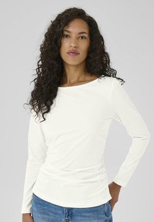 White long-sleeve top with a boat neckline and gathered detail on the side, made of smooth fabric, paired with blue jeans.