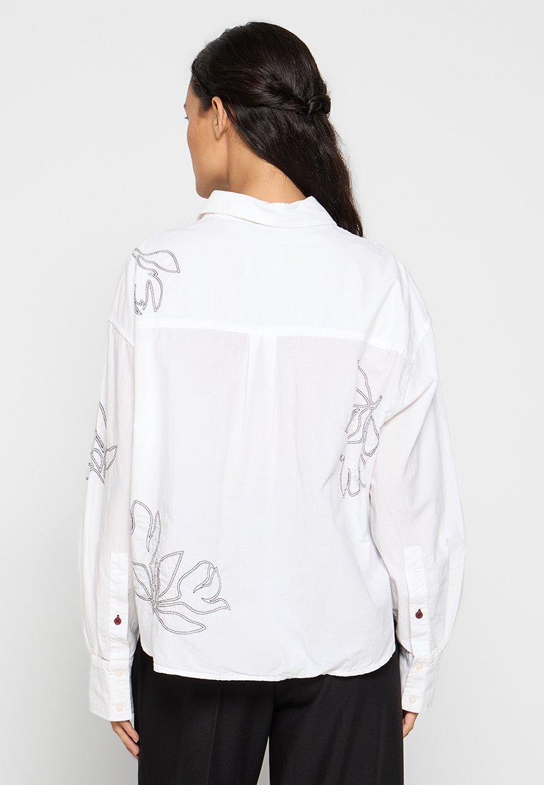 Witte geborduurde blouse met lange mouwen. Beschikt over bloemenontwerpen in grijs op de schouders en rug, knopenmanchetten en een klassieke kraag.