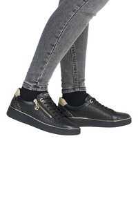 Baskets noires avec un dessus texturé présentant un motif géométrique, des accents dorés, des détails zippés et une semelle en caoutchouc, associées à un jean skinny gris.