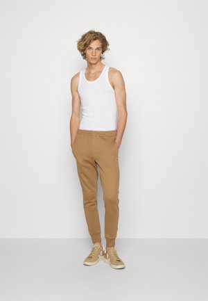 Brown sweatpants, elastic waistband, and cuffs; white tank top. Model wearing beige sneakers with white accents. Plain background.