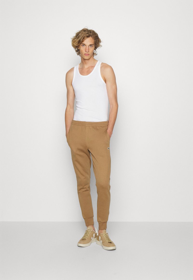 Pantaloni de trening maro, cu talie elastică și mansete; tricou alb fără mâneci. Modelul poartă adidași bej cu accente albe. Fundal simplu.