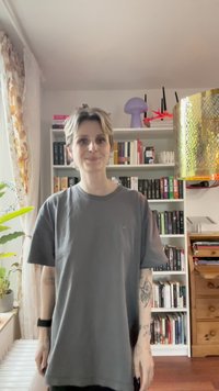 T-shirt en coton gris à manches courtes et coupe décontractée, avec un petit logo vert sur la poitrine. L'arrière-plan comprend des étagères de livres et de la décoration.