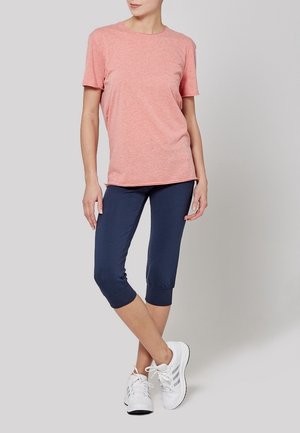 Woman wearing a pink short-sleeve T-shirt, navy capri pants, and white sneakers, standing with legs crossed against a plain background.