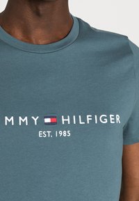 Tyrkysové bavlněné tričko s kulatým výstřihem a krátkými rukávy. Obsahuje bílé vyšívané jméno značky a logo s modrým, červeným a bílým akcentem.