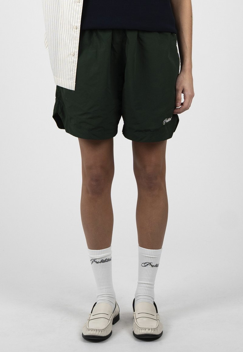 Personne portant un short vert foncé, des chaussettes blanches avec un texte noir et des mocassins crème, debout devant un fond clair uni.