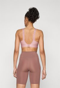 Lys rosa sports-bh med brede stropper, detaljert logo og lav rygg. Kombinert med høytlivs sykkelshorts i rustfarget med sidelommer.
