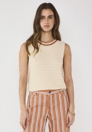 Jeune femme aux cheveux longs portant un haut sans manches en maille crème et un pantalon taille haute marron à rayures verticales blanches, debout devant un fond uni.